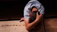 Ein Junge verschränkt die Arme über seinem auf einen Holztisch gelegten Kopf und versteckt damit sein Gesicht.