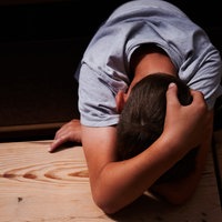 Ein Junge verschränkt die Arme über seinem auf einen Holztisch gelegten Kopf und versteckt damit sein Gesicht.
