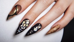Lackierte, verlängerte Nägel in Schwarz-Gold, besetzt mit Edelsteinen.