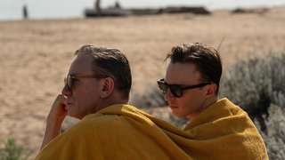In dieser Szene aus dem Film „Queer“ von Luca Guadagnino sitzen Daniel Craig und Drew Starkey in ihren Rollen mit nassen Haaren und Sonnenbrillen nebeneinander – gemeinsam tragen sie ein Handtuch über den Schultern.