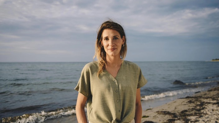 Eine Porträtaufnahme von Rina Schmeller zeigt sie mit freundlichem Lächeln an einem Strand.