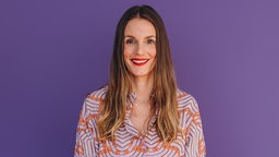 Die Porträtaufnahme von Sara Buschmann zeigt sie mit einem freundlichen Lächeln vor einem lila Hintergrund.