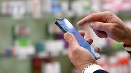 Symbolbild zur Sicherheit von Apotheken-Apps: Ein Mann hält sein Handy in der Hand, im Hintergrund die Regale einer Apotheke