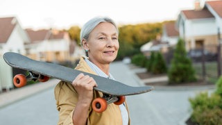 Symbolbild: Eine Frau mit grauem Haar trägt lässig ein Skateboard über ihrer Schulter udn lächelt dabei.