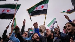 Menschen feiern und schwenken syrische Flaggen, während sie in Damaskus, Syrien, am Montag, dem 8. Dezember 2025, auf eine Parade der neuen syrischen Armee zum ersten Jahrestag der Absetzung des Bashar-al-Assad-Regimes warten.
