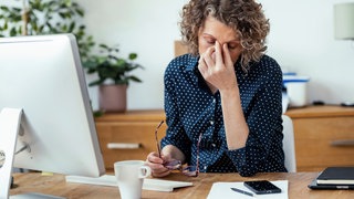 Eine Frau massiert sich mit Daumen und Zeigefinger den Punkt an der Stirn direkt über der Nase, während sie vor einem Computerbildschirm sitzt.