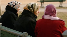Ältere Muslima mit Kopftuch sitzen auf einer Bank auf einem Platz. 