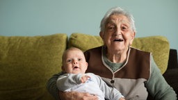 Eine glückliche Urgroßmutter mit grauem kurzem Haar sitzt auf einem Sofa mit ihren Urenkel im Arm. 