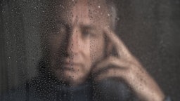 Hinter einer mit Tropfen bedeckten Glasscheibe ist verschwommen das Gesicht eines nachdenklichen Mannes zu erkennen.