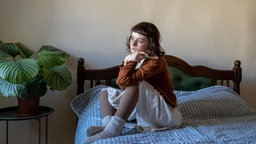 Auf dem Bild ist eine melancholische Frau zu sehen, die auf dem Bett sitzt und ins Leere blickt.