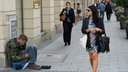 Ein Obdachloser sitzt in einer Fußgängerzone, während wohlhabend gekleidete Frauen dort einkaufen gehen.