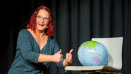 Die Kabarettistin Anny Hartmann auf der Bühne, sie hält lachend beide Daumen nach oben. Neben ihr steht ein Stuhl, auf dem ein Globus liegt.