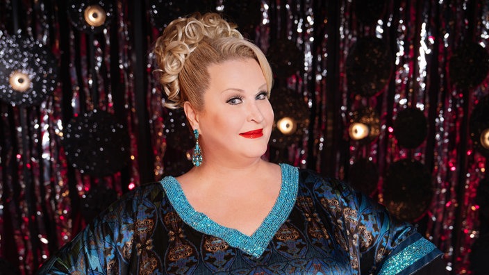 Daphne de Luxe vor einem glitzernden Vorhang.