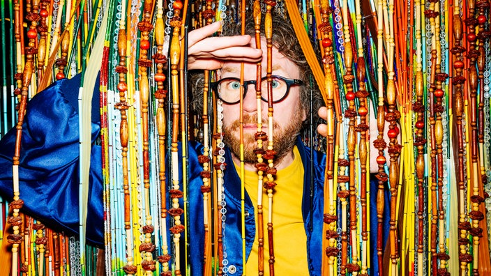 Friedemann Weise schaut in strahlende blauer Jacke gekleidet heimlich durch einen bunten Perlenvorhang in die Kamera, Foto: Frederike Wetzels