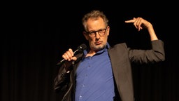 Severin Groebner hält ein Mikrofon und steht einbeinig auf der Bühne. Er zeigt mit dem eigenen Zeigefinger auf sich selbst. Foto: Dominic Reichenbach