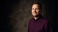 Der Comedian Jakob Schwerdtfeger steht verschmitzt lächelnd vor einem dunklen Hintergrund