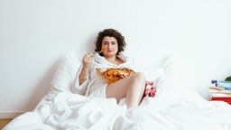 Comedienne Matilde Keizer sitzt mit weißem Bademantel im weiß bezogenen Bett und isst mit einer Gabel Lasagne aus einer Auflaufform