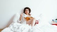 Comedienne Matilde Keizer sitzt mit weißem Bademantel im weiß bezogenen Bett und isst mit einer Gabel Lasagne aus einer Auflaufform
