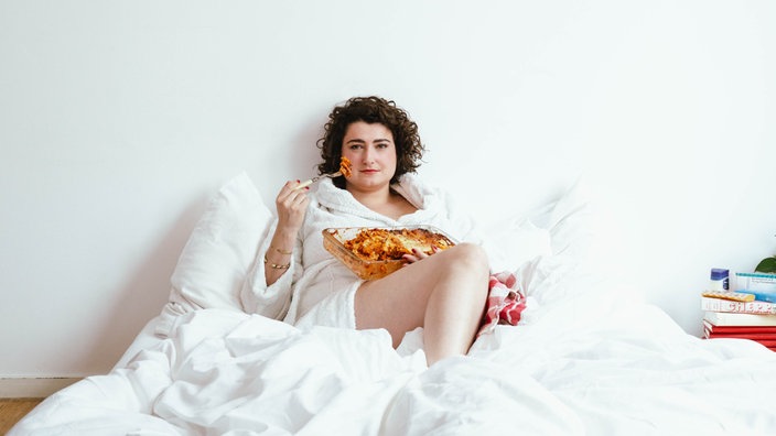 Comedienne Matilde Keizer sitzt mit weißem Bademantel im weiß bezogenen Bett und isst mit einer Gabel Lasagne aus einer Auflaufform