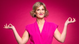 Die Comedienne Vera Deckers steht vor einem rötlich-pinken Hintergrund, trägt ein pinkes Kostüm und lächelt in die Kamera.