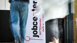  Personen gehen am Logo vom Jobcenter vorbei.