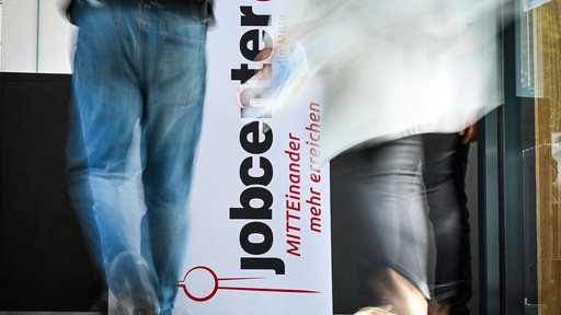  Personen gehen am Logo vom Jobcenter vorbei.