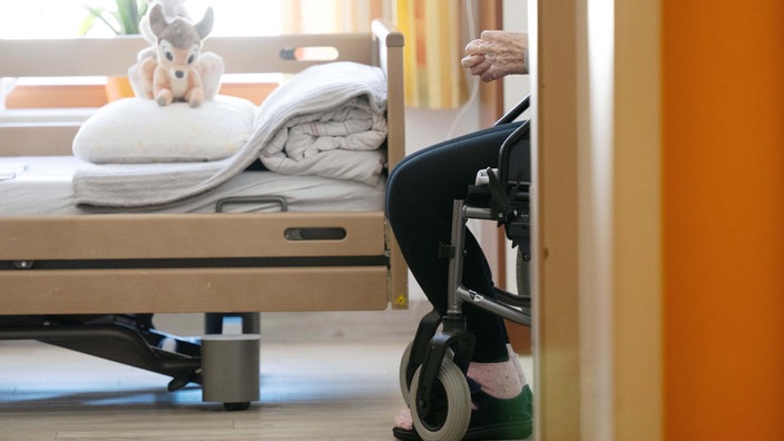 Ein Bewohner sitzt in einem Altenheim in seinem Zimmer in einem Rollstuhl. Auf dem gemachten Bett liegt ein Stofftier.