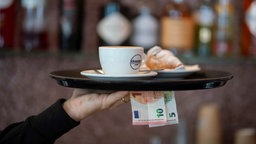 Nahaufnahme eines Arms eines Kellners oder einer Kellnerin in einem schwarzen Pullover, der ein Tablett mit einer Tasse und einem Croissant hält – außerdem befinden sich ein 5 Euro Schein und ein 10 Euro Schein in der Hand unter dem Tablett. 
