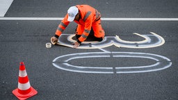 Ein Mann in orangener Arbeitsuniform klebt auf einer Straße mit Klebeband die Zahl "30" auf, um sie anschließend dort aufmalen zu können.