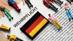 Neben einer Deutschlandflagge liegen mehrere Figuren, die Bürger darstellen, auf einem Papier mit der Überschrift "Wehrpflicht".