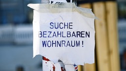An einem Ampel- oder Laternenpfahl hängt ein weißer Zettel mit der blauen Aufschrift "Suche bezahlbaren Wohnraum!".