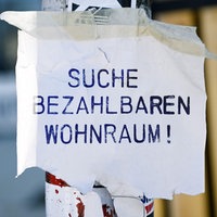 An einem Ampel- oder Laternenpfahl hängt ein weißer Zettel mit der blauen Aufschrift "Suche bezahlbaren Wohnraum!".