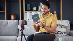 Ein junger Männ hält lächelnd eine Bibel in eine Kamera. 