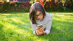 Mädchen liegt auf einer Wiese und schaut in ein Smartphone. 