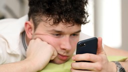 Teenager schaut im Bett gelangweilt auf sein Smartphone.  