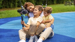 Mutter und zwei Söhne sitzen auf einem  Spielplatz und erstellen  Videoinhalte mit einem Smartphone.