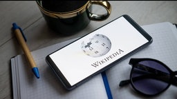 Notizblock, Stift und Lesebrille liegen neben einem Smartphone mit der Aufschrift "Wikipedia".