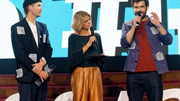 Die Autoren Hans Block (l), Moritz Riesewieck (r) stehen bei Verleihung der Grimme-Online-Awards mit Moderatorin Anja Backhaus mit ihren Preis in der Kategorie "Sonderpreis Künstliche Intelligenz" auf der Bühne.