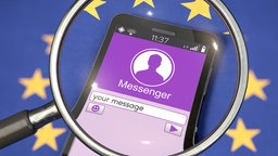 Auf einer Europaflagge liegt ein Smartphone. Eine Lupe vergrößert den Bildschirm, der eine Messenger-App zeigt.