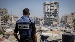 Ein Journalist mit schusssicherer Presse-Weste steht in Gaza mit dem Rücken zur Kamera und blickt über eine Zeltstadt vor zerstörten Gebäuden.