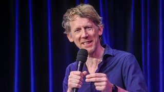Christian Schulte-Loh auf der Bühne beim WDR5 Kabarettfest in Köln vom 21.09.2025, hält Mikro in der Hand und spricht lächelnd zum Publikum 