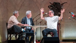Das Kabarett-Trio "Wir Staatskünstler" sitzt beim Salzburger Stier 2026 an einem Tisch auf der Bühne