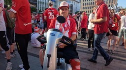 Ein RWE-Fan im Trikot sitzt in einem E-Rollator in einer Menschenmenge