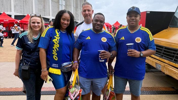 Zwei Fußballfans aus Hagen stehen gemeinsam mit Fans aus Curaçao draußen vor einem Stadion, sie tragen blaue Fußballtrikots und halten Getränke in der Hand.
