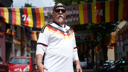 Achim Klimmeck in einem Deutschland‑Fußballtrikot, schwarzer Kappe und Sonnenbrille. Im Hintergrund hängen dutzende Deutschlandflaggen und ein Mannschaftsbild.