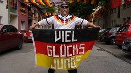 Achim Klimmeck in einem Deutschland‑Trikot, er hält eine große schwarz‑rot‑goldene Flagge, auf der in weißen Buchstaben "Viel Glücks Jungs" steht. Im Hintergrund eine Straße mit Deutschland‑Flaggen als Dekoration.