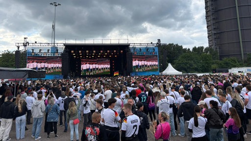 In Oberhausen können bis zu 40.000 Menschen das erste Spiel der deutschen Nationalmannschaft bei der WM 2026 sehen. Auf großen Leinwänden ist zu sehen, wie die deutschen Spieler die Nationalhymne singen. Der Platz vor den Leinwänden ist mit Fans gefüllt.