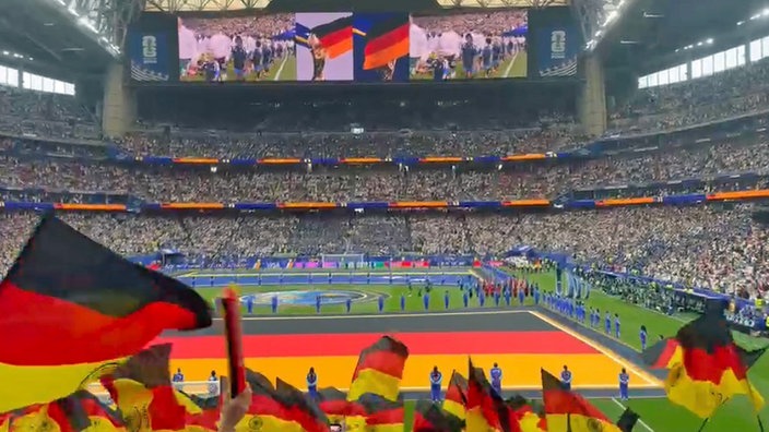Fans schwenken Deutschlandfahnen in einem Stadion, während auf dem Spielfeld vor großer Videoleinwand die Einlaufzeremonie stattfindet.