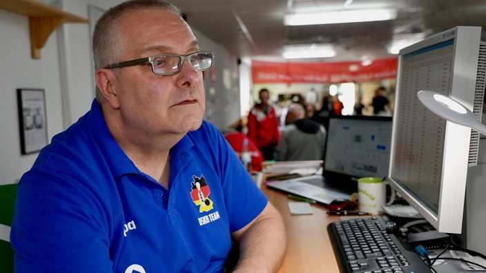 Ein mittelalter Herr mit Halbglatze, einer schmalen eckigen Brille und einem leuchtend blauen T-Shirt.
