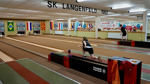 Man sieht eine Kegelbahn, eine sportliche Frau ist gerade am Kegeln.